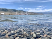 Clear, shallow water with smooth pebbles visible beneath the surface. In the background, a line of wind turbines is situated on a grassy landscape, with mountains partially covered in snow under a blue sky with scattered clouds.