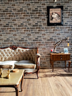 An elegant living room features a vintage-style sofa with plush cushions placed on a wooden floor. The wall is adorned with rustic brick wallpaper, enhancing the room's cozy and classic ambiance. A framed poster with text and an image of a tattooed individual is mounted on the wall. Beside the sofa, a stylish wooden side table holds a decorative branch with a hanging light bulb, along with a small candle and picture frame. In front of the sofa, a coffee table with golden figurines adds a touch of luxury.