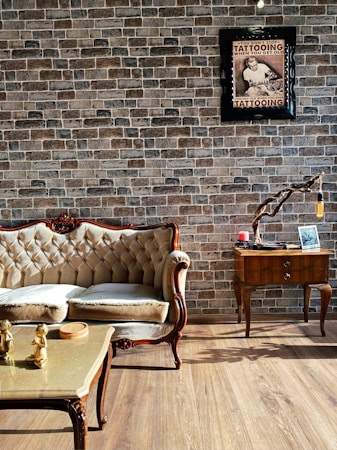 An elegant living room features a vintage-style sofa with plush cushions placed on a wooden floor. The wall is adorned with rustic brick wallpaper, enhancing the room's cozy and classic ambiance. A framed poster with text and an image of a tattooed individual is mounted on the wall. Beside the sofa, a stylish wooden side table holds a decorative branch with a hanging light bulb, along with a small candle and picture frame. In front of the sofa, a coffee table with golden figurines adds a touch of luxury.