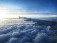 An airplane soaring above fluffy white clouds with the sun setting on the horizon.