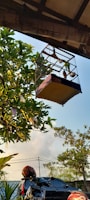 A bird cage is hanging outdoors under a roof, surrounded by green leafy trees. In the background, there are power lines and a partly cloudy sky. Below the bird cage, there is a parked motorbike with a helmet placed on it.