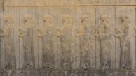 A stone relief depicting a line of ancient figures wearing robes and headdresses, holding staffs. The relief features intricate carvings with a floral pattern border above and below the figures.