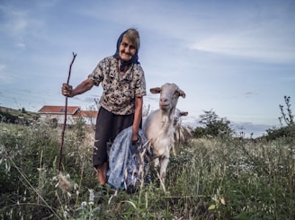An elderly woman smiling while holding a shopping basket with a friendly companion assisting her.