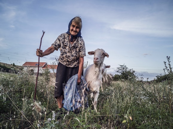 An elderly woman wearing a floral shirt and a headscarf is holding a walking stick and standing next to a goat in a grassy field. She is smiling and holding a bag in one hand. The background includes a house with a red roof and some trees under a partly cloudy sky.