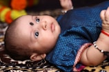 An infant lies on a patterned surface, looking upwards with wide eyes. The baby is wearing a blue outfit with intricate designs and has a black dot on the forehead. Bright colors, such as orange and yellow, are slightly visible in the background.