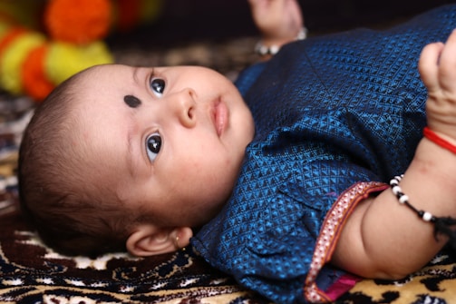 An infant lies on a patterned surface, looking upwards with wide eyes. The baby is wearing a blue outfit with intricate designs and has a black dot on the forehead. Bright colors, such as orange and yellow, are slightly visible in the background.