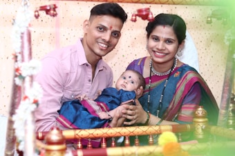 A joyful family picture featuring a couple holding a baby, surrounded by a decorated setting with flowers and colorful drapes. The parents are smiling warmly, and the baby is dressed in vibrant traditional attire.