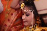 A woman adorned in traditional attire with intricate jewelry, including a nose ring and decorative headpiece. Her hair is embellished with yellow flowers and she appears to be in a ceremonial setting, indicated by the presence of vibrant orange and gold fabrics in the background.