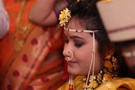 A woman adorned in traditional attire with intricate jewelry, including a nose ring and decorative headpiece. Her hair is embellished with yellow flowers and she appears to be in a ceremonial setting, indicated by the presence of vibrant orange and gold fabrics in the background.