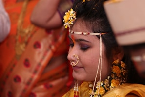 A woman adorned in traditional attire with intricate jewelry, including a nose ring and decorative headpiece. Her hair is embellished with yellow flowers and she appears to be in a ceremonial setting, indicated by the presence of vibrant orange and gold fabrics in the background.