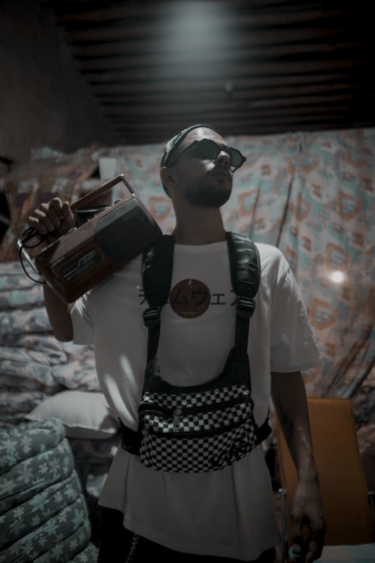 A person wearing sunglasses and a white shirt with a graphic, holding an old-fashioned radio on their shoulder. They have a checkered crossbody bag and stand in a room with patterned bedding and curtains.