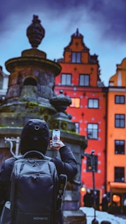 A person wearing a black hooded jacket and backpack is taking a photo with a smartphone in front of an old fountain. In the background, there are colorful historic buildings with ornate facades, including a prominent red building with large windows. The sky appears cloudy, adding a dramatic effect to the scene.