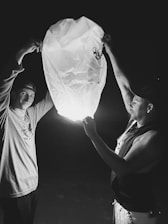Two people are holding a lit lantern in a dark setting, creating a warm glow that illuminates their faces. They appear engaged and focused on the lantern as they carefully let it rise into the sky.