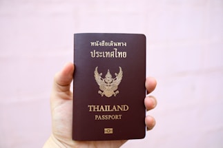 A hand is holding a brown passport with an emblem and text on the cover that indicates it is a Thailand passport. The text is in both Thai and English, and the background is plain and light-colored.