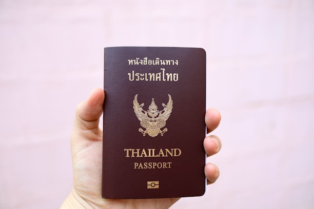 A hand is holding a brown passport with an emblem and text on the cover that indicates it is a Thailand passport. The text is in both Thai and English, and the background is plain and light-colored.