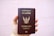 A hand is holding a brown passport with an emblem and text on the cover that indicates it is a Thailand passport. The text is in both Thai and English, and the background is plain and light-colored.