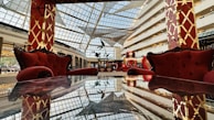 A luxurious interior space featuring plush red armchairs and sofas against a backdrop of a large glass ceiling, allowing natural light to illuminate the area. The furniture is placed around a reflective table surface, enhancing the opulent and grand ambiance. The decor includes elaborate golden patterns on red pillars.