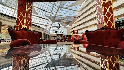 A luxurious interior space featuring plush red armchairs and sofas against a backdrop of a large glass ceiling, allowing natural light to illuminate the area. The furniture is placed around a reflective table surface, enhancing the opulent and grand ambiance. The decor includes elaborate golden patterns on red pillars.