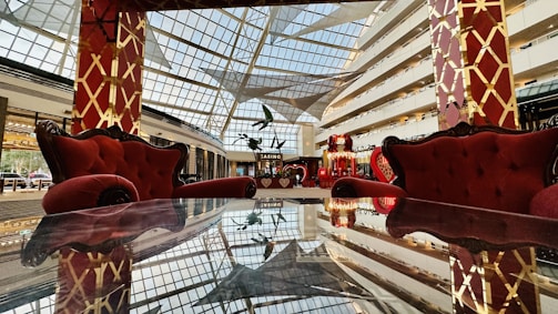 A luxurious interior space featuring plush red armchairs and sofas against a backdrop of a large glass ceiling, allowing natural light to illuminate the area. The furniture is placed around a reflective table surface, enhancing the opulent and grand ambiance. The decor includes elaborate golden patterns on red pillars.