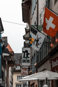 A narrow street lined with buildings featuring signs and flags. Most prominent is the red flag with a white cross, representing Switzerland. Other signs include an image of a person in a black hat and various business signs. The architecture is traditional European with sloping roofs and shutters.