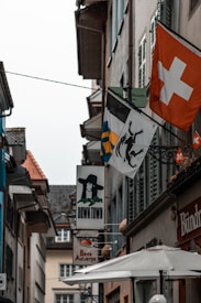A narrow street lined with buildings featuring signs and flags. Most prominent is the red flag with a white cross, representing Switzerland. Other signs include an image of a person in a black hat and various business signs. The architecture is traditional European with sloping roofs and shutters.