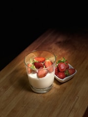 A glass filled with white yogurt topped with fresh strawberries is placed on a wooden surface. Beside the glass, a small white dish holds several whole strawberries. The scene is illuminated with soft lighting, creating a cozy and inviting atmosphere.