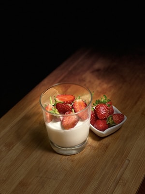 A glass filled with white yogurt topped with fresh strawberries is placed on a wooden surface. Beside the glass, a small white dish holds several whole strawberries. The scene is illuminated with soft lighting, creating a cozy and inviting atmosphere.