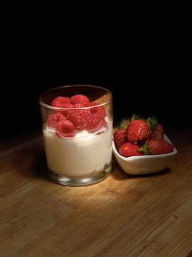 A glass filled with creamy white yogurt is topped with bright red raspberries, set next to a white dish containing fresh strawberries. The setup is placed on a wooden surface with a dark background, highlighted by soft lighting that enhances the vivid colors of the fruits.