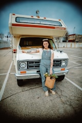 A person stands in front of a vintage RV parked in a nearly empty parking lot. The person is wearing overalls, a white shirt, and has a bag filled with plants in one hand. The RV has a classic design with a roof-mounted light and the word 'Coachmen' on it. The background shows some buildings and trees under a cloudy sky.