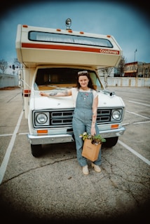 A person stands in front of a vintage RV parked in a nearly empty parking lot. The person is wearing overalls, a white shirt, and has a bag filled with plants in one hand. The RV has a classic design with a roof-mounted light and the word 'Coachmen' on it. The background shows some buildings and trees under a cloudy sky.