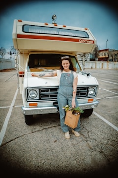 A person stands in front of a vintage RV parked in a nearly empty parking lot. The person is wearing overalls, a white shirt, and has a bag filled with plants in one hand. The RV has a classic design with a roof-mounted light and the word 'Coachmen' on it. The background shows some buildings and trees under a cloudy sky.