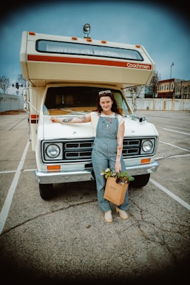 A person stands in front of a vintage RV parked in a nearly empty parking lot. The person is wearing overalls, a white shirt, and has a bag filled with plants in one hand. The RV has a classic design with a roof-mounted light and the word 'Coachmen' on it. The background shows some buildings and trees under a cloudy sky.