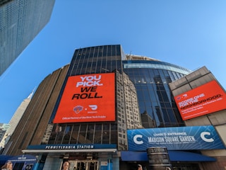 A cityscape image featuring the entrance of Madison Square Garden with large digital advertisements on the building facade. The advertisements promote a partnership with the New York Knicks and DoorDash, featuring bold typography and vibrant orange backgrounds. The iconic Empire State Building is visible in the background, and the clear blue sky adds to the urban setting. The signage for Pennsylvania Station is prominently displayed below.