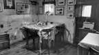 A rustic and vintage kitchen or dining room with a wooden table covered by a polka-dot tablecloth. Two chairs are positioned around the table. On the table, there is a basket with fruit and a bottle. The walls are adorned with various framed pictures and the paint is somewhat worn. A small toaster is placed on a wooden sideboard near the door, and there is minimal natural light coming through a small window.