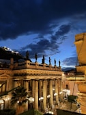 A historic theater with classical architecture, featuring tall columns and sculptures along the rooftop. It is illuminated by street lamps, with a dramatic nighttime sky above, filled with dark clouds against a fading blue backdrop.