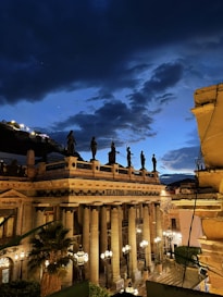 A historic theater with classical architecture, featuring tall columns and sculptures along the rooftop. It is illuminated by street lamps, with a dramatic nighttime sky above, filled with dark clouds against a fading blue backdrop.