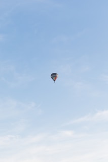 A solitary hot air balloon floats against a vast expanse of light blue sky. The balloon has a colorful pattern and appears to be high in the sky, conveying a sense of freedom and adventure.