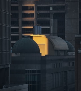 A modern architectural scene featuring a building with distinct dome-shaped structures and industrial design. The image captures a warm, soft light highlighting a portion of the façade, contrasting with the shadowed areas of the building. The structure is surrounded by tall, dark buildings in the background, creating a dramatic play of light and shadow.
