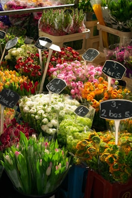 A vibrant array of flowers in a market setting, featuring bouquets of various colors such as red, pink, white, orange, and green. Each bouquet is placed in a bucket and labeled with price tags in Euros, indicating a busy and colorful floral stall.