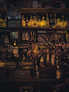 A dimly lit vintage bar shelf filled with an assortment of liquor bottles, books, and decorative items. The bottles, which are mostly whiskey, are neatly arranged on the top shelf and illuminated by a warm, soft light. Various memorabilia, including old photographs, framed pictures, and musical instruments, such as a saxophone, are scattered around the shelves and surfaces. The setting conveys a sense of nostalgia and a cozy, intimate atmosphere.