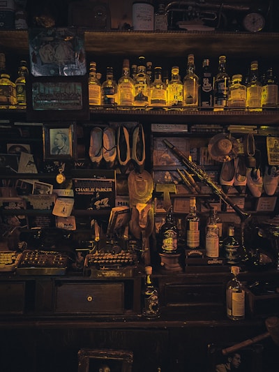 A dimly lit vintage bar shelf filled with an assortment of liquor bottles, books, and decorative items. The bottles, which are mostly whiskey, are neatly arranged on the top shelf and illuminated by a warm, soft light. Various memorabilia, including old photographs, framed pictures, and musical instruments, such as a saxophone, are scattered around the shelves and surfaces. The setting conveys a sense of nostalgia and a cozy, intimate atmosphere.