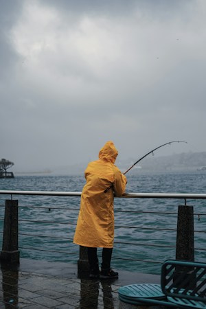 Angler in a yellow hooded rain jacket leaning on a waterfront railing with a bent rod, gray water and a misty shoreline beyond