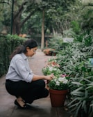 A person crouches down to tend to potted flowers along a garden path. The area is lush with greenery and flowering plants, creating a serene and natural environment.