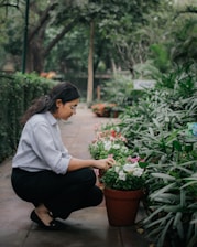 A person crouches down to tend to potted flowers along a garden path. The area is lush with greenery and flowering plants, creating a serene and natural environment.