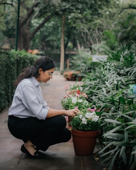 A person crouches down to tend to potted flowers along a garden path. The area is lush with greenery and flowering plants, creating a serene and natural environment.