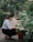 A person crouches down to tend to potted flowers along a garden path. The area is lush with greenery and flowering plants, creating a serene and natural environment.