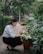 A person crouches down to tend to potted flowers along a garden path. The area is lush with greenery and flowering plants, creating a serene and natural environment.