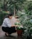 A person crouches down to tend to potted flowers along a garden path. The area is lush with greenery and flowering plants, creating a serene and natural environment.