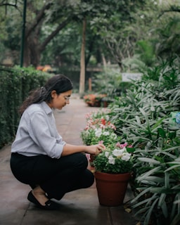 A person crouches down to tend to potted flowers along a garden path. The area is lush with greenery and flowering plants, creating a serene and natural environment.