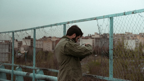 A person wearing a green jacket is standing near a chain-link fence with barbed wire on top. The scene appears to be on a bridge or overpass with a cityscape in the background. The mood is somewhat overcast, with muted colors dominating the environment.